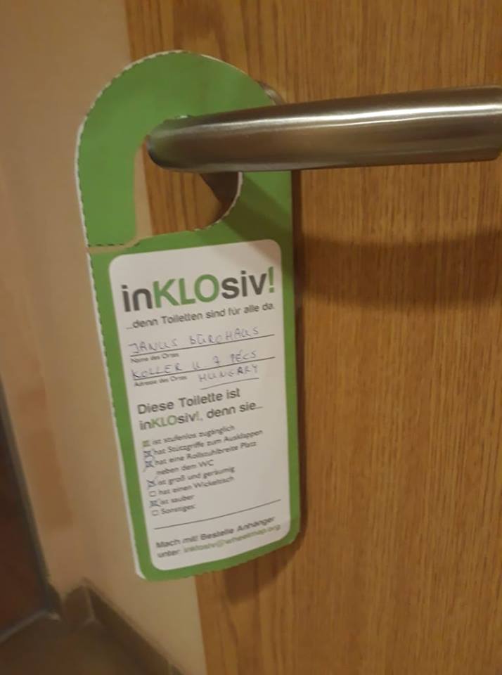 A kilincsen lóg az általunk odaakasztott papír fülecske inKLOsiv felirattal, és az akadálymentes WC jellemzőivel németül