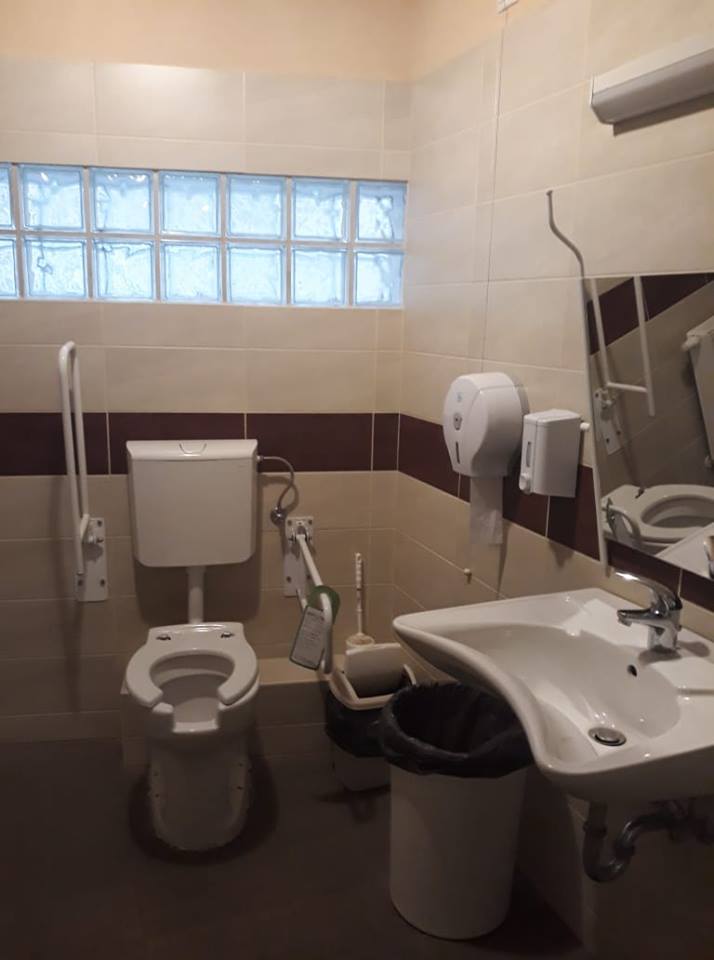 Az irodaház akadálymentes WC-je, szemben a WC kétoldalán kapaszkodókkal. Jobb oldalon a kézmosó, mellette baloldalt a falon a szappantartó és WC papír tartó.
