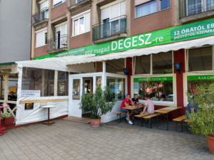 Kintről a terasz az utcáról nézve. A teraszon sörpadok és asztalok egy napernyő alatt. A bejárat szintén fedett kintről.