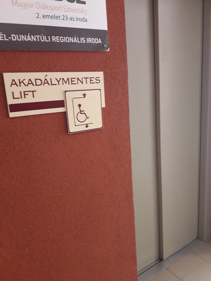 Felirat a falon az akadálymentes WC-t jelezve lift mellett. Azt mutatja, hogy akadálymentes WC-t a lifttel fölmenve egy emelettel feljebb találunk. Braille felirat nincs rajta.