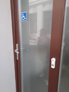 A WC bejárata az utcáról, befelé nyíló átláthatatlan üvegajtóval. Az ajtó jobboldalán kis csengőt kell nyomni ahhoz, hogy kinyissák.