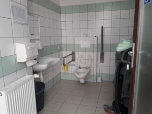 A WC újabb képen, minden oda nem illő dolgot kipakoltak. Belépve szemben balra a falon radiátor, mellette a kézmosó fali papírtörlő tartóval és szappanadagolóval. A szemben lévő falon balra a WC szabályos kapaszkodókkal, vészcsengővel balra a falon. Jobboldalt még mindig valami takarítógép parkol az akadálymentes WC-ben.