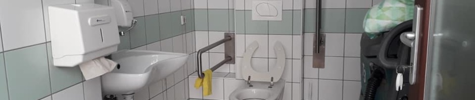 A WC újabb képen, minden oda nem illő dolgot kipakoltak. Belépve szemben balra a falon radiátor, mellette a kézmosó fali papírtörlő tartóval és szappanadagolóval. A szemben lévő falon balra a WC szabályos kapaszkodókkal, vészcsengővel balra a falon. Jobboldalt még mindig valami takarítógép parkol az akadálymentes WC-ben.