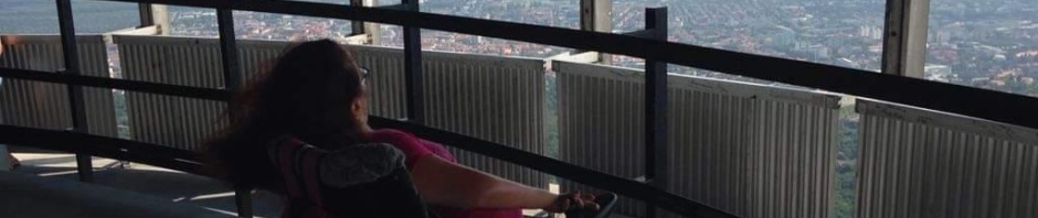 A TV torony teraszán áll kerekesszékes tagunk és gyönyörködik az elétáruló város panorámájában. A terasz nyitott, a fenti részbe lehet kerekesszékkel kijutni, a lenti részhez lépcső vezet.