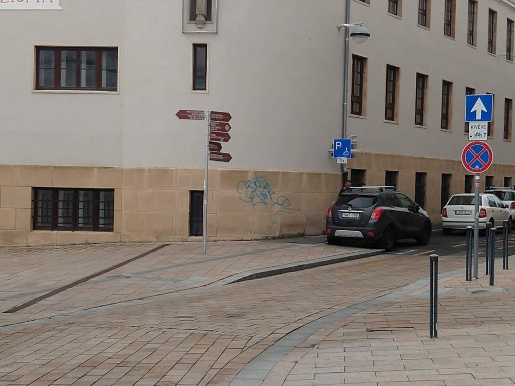 A Széchenyi tér fölső részén a Janus Pannonius utca sarkán a Nagy Lajos gimnázium falán látható az akadálymentes parkolókat jelző tábla.