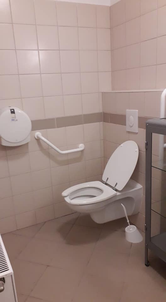 Csempézett oldalú WC, fehér csempével, középen szürke csempesorrel, szürke járólappal. Az egyik oldalt falra szerelhető müanyaggal bevont kapaszkodóval.