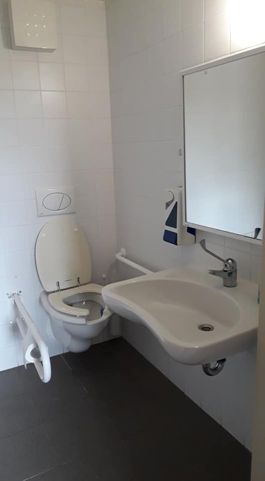 Illemhely fehér csempével, szürke járólappal. A WC felett gomb az öblítéshez, mellette kétoldalt kapaszkodó. A kép jobboldalán mosdókagyló, tükörrel, jobboldalt szappanadagolóval.