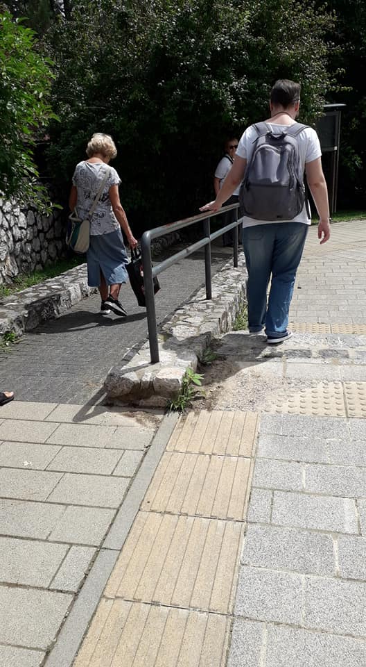 A tér szélén lévő szürke térköves járda, szélén a sárga vakvezzetősávokkal, mellette a fekete vaskorláttal ellátott fekete köves rámpa. A korlát jobboldalán egy férfi a baloldalán egy nő megy lefelé, a kép készítőjének háttal.