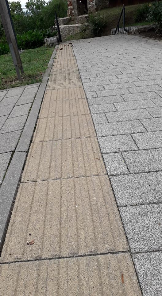 A Tettye tér tetejéről lefelé készült a kép, felül belóg egy részlet a romokból. A kép fókuszában a szürke térköves járda, szélén a sárga vakvezető sávokkal.