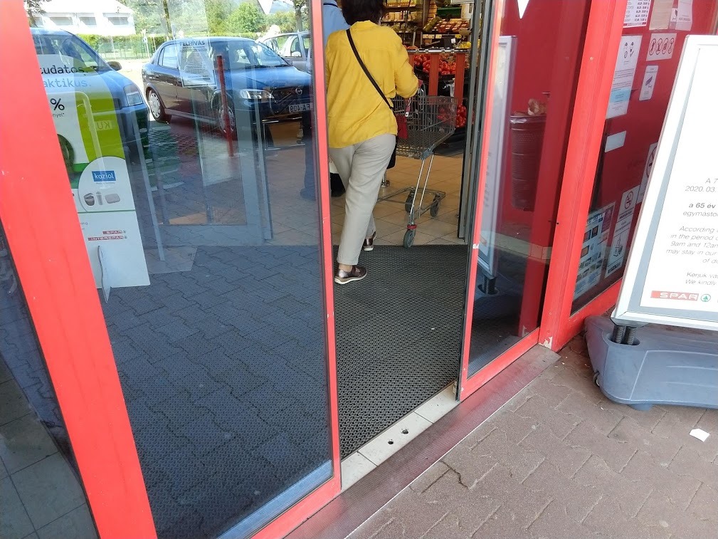 A Spar fotocellás ajtaja félig nyitva, tehát mozgásban, kívülről fotózva. Az áruházba egy sárga inges nő megy befelé, bevásárlókocsit tolva.
