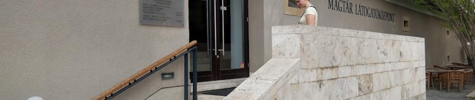 A Magtár Látogatóközpont bejárata látható kívülről. Az ajtó mészkőből készült lépcsőn közelíthető meg, a lépcső külső oldalán masszív párkány, a fal felőli oldalon fémből és fából készített korlát látható. Az épület fala szürke, a háttérben láthatjuk a kávézó teraszát. A lépcső tetején egy fiatal nő áll.