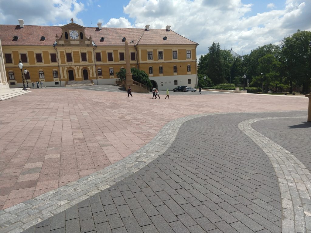 A képen a Dóm tér látható felülről, a Bazilika jobb oldaláról a tér közepe felé fotózva. A tér üres, szélén szürke, középen vörös térkővel. Szemben a Camera Sepolcrale sárga épülete látható.