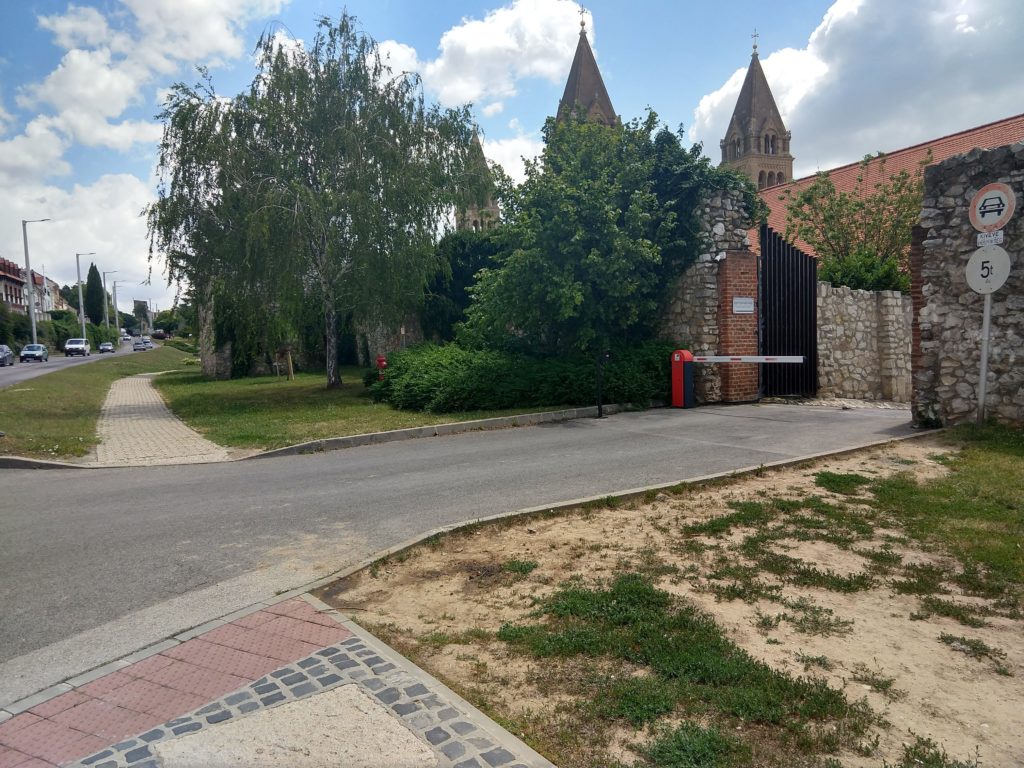 A Magtár Látogatóközpont Aradi vértanúk útjára nyíló kocsibejárója látható kívülről. Az út kétoldalán a járda szélén a járda végét jelző sávok. Háttérben láthatóak a bazilika tornyai előtte a magas kőfal mentén zöldellő fák.A Magtár Látogatóközpont Aradi vértanúk útjára nyíló kocsibejárója látható kívülről. Az út kétoldalán a járda szélén a járda végét jelző sávok. Háttérben láthatóak a bazilika tornyai előtte a magas kőfal mentén zöldellő fák.