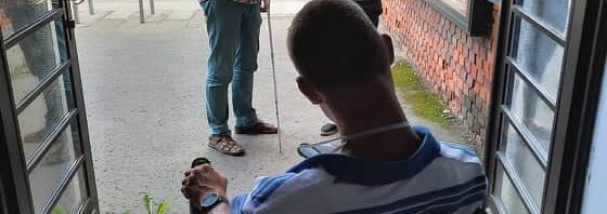 Az uránia mozi bejárata az épületből kifelé fotózva látható. Az épületben a fénykép készítőjének háttal egy kerekesszékes férfi csíkos pólóban, vele szemben a mozi előtti kis aszfaltozott téren egy feketepólós nő és egy vak férfi kockás ingben és farmerban.