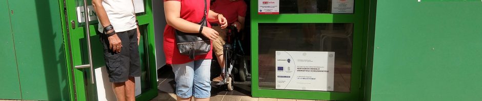 A Csontváry rendelő bejáratával állunk szemben. Az épület zöld színű. A képen szerepel 3 ember. Egy férfi támasztja az ajtót, mögötte egy kerekesszékes férfi jön ki az ajtón. Az úr előtt egy hölgy áll.