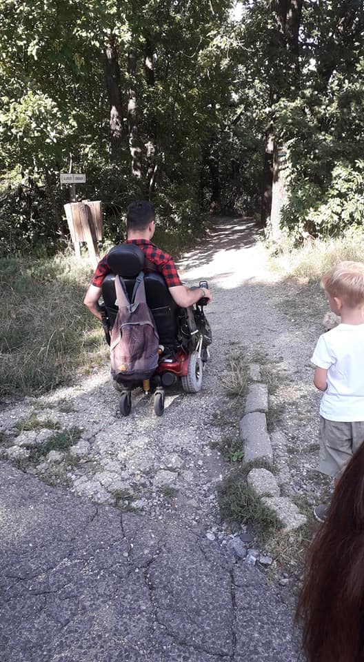 Egy kerekesszékes férfi gurul a kavicsos úton, ami egy erdőbe vezet. Piros kockás ing van rajta, a kerekesszék hátán lóg egy barna hátizsák. Mögötte áll egy kisfiú, fehér pólóban és szürke rövidnadrágban.