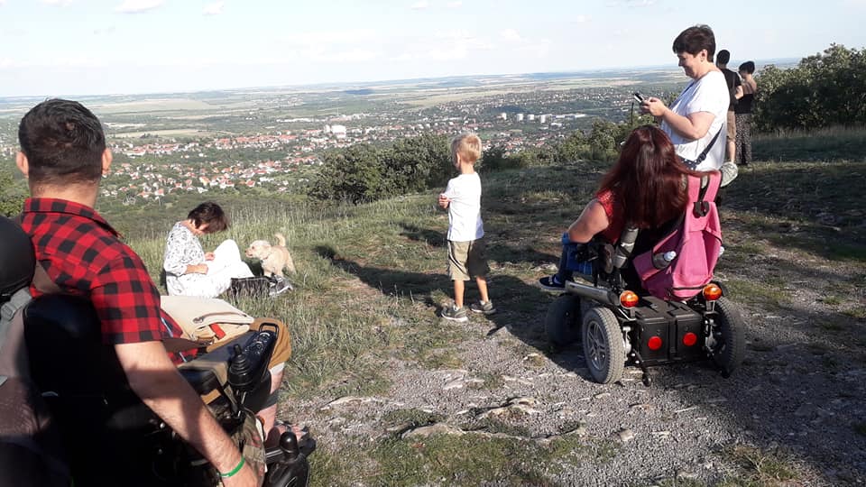 Flóra pihenő. Szép az idő, süt a nap. Hét ember szerepel a képen, akik épp a tájat nézik. Közülük ketten, egy férfi és egy nő motoros kerekesszékben ül. Szerepen a képen egy kiskutya is, barna szőrű, kis termetű.
