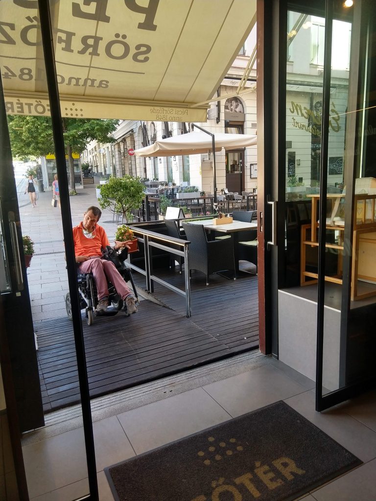 A főtér bisztró bejárata látható. A bejárattal szemben egy kerekesszékes férfi található, aki épp jön fel a rámpán.