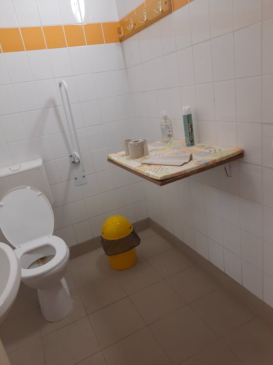 Egy wc található a képen. Velünk szemben található a wc, mellette egy kapaszkodóval. A jobb oldali falon helyet kapott egy pelenkázó, mellette egy kukával. Bal oldalon található a mosdókagyló.