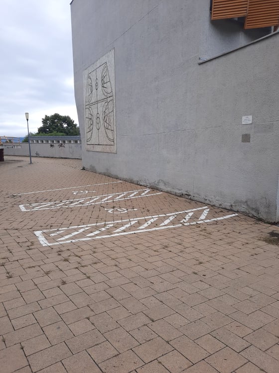 Egy parkolóban vagyunk. A parkoló burkolata barna térkő. Egy nagy szürke épület van a parkoló jobb szélén. Két mozgássérült parkolóhely van felfestve a térkőre.