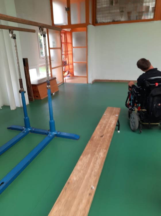 Egy tornateremben vagyunk. Zöld gumi borítású a padló. A teremben van egy fa pad. A pad mellett egy kerekesszékes férfi szerepel.