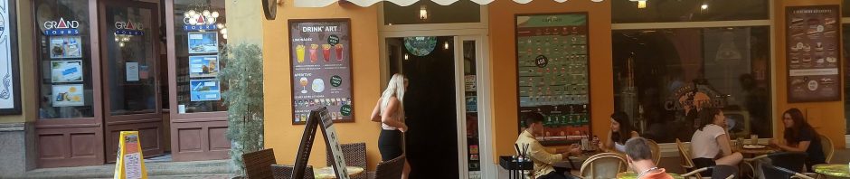 A Cefe Frei kiülős helyén vagyunk. Ez egy sárga épület napernyővel ellátva. Az épület falain pedig a kávékülönlegességek találhatóak. A főbejáratnál egy kerekesszékes úriember szeretne bejutni a kávézóba. A bejáratnál van egy kisebb patka és még egy küszöb is van rajta, viszont egy erősebb mozgáskorlátozott vagy segítséggel be lehet jutni. Bal oldalt a Grand Tours Kft. található.