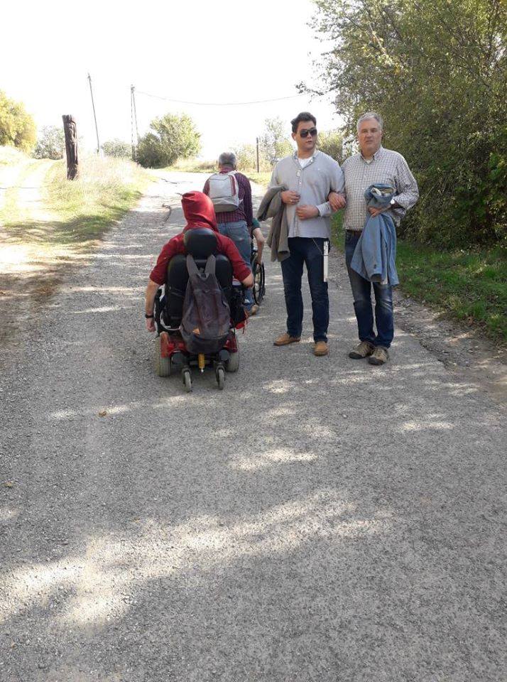 Egy rossz állapotban lévő beton úton látható egy csoport ember, pillanatképe. Elöl egy idősebb, szemüveges uriember egyenesen a kamerába néz, zsebre tett kézzel. Mögötte egy kicsit távolabb pár ember, két kerekesszékes uriemberrel. Mögöttük a tájat eltakaró, zöld növényzet található.