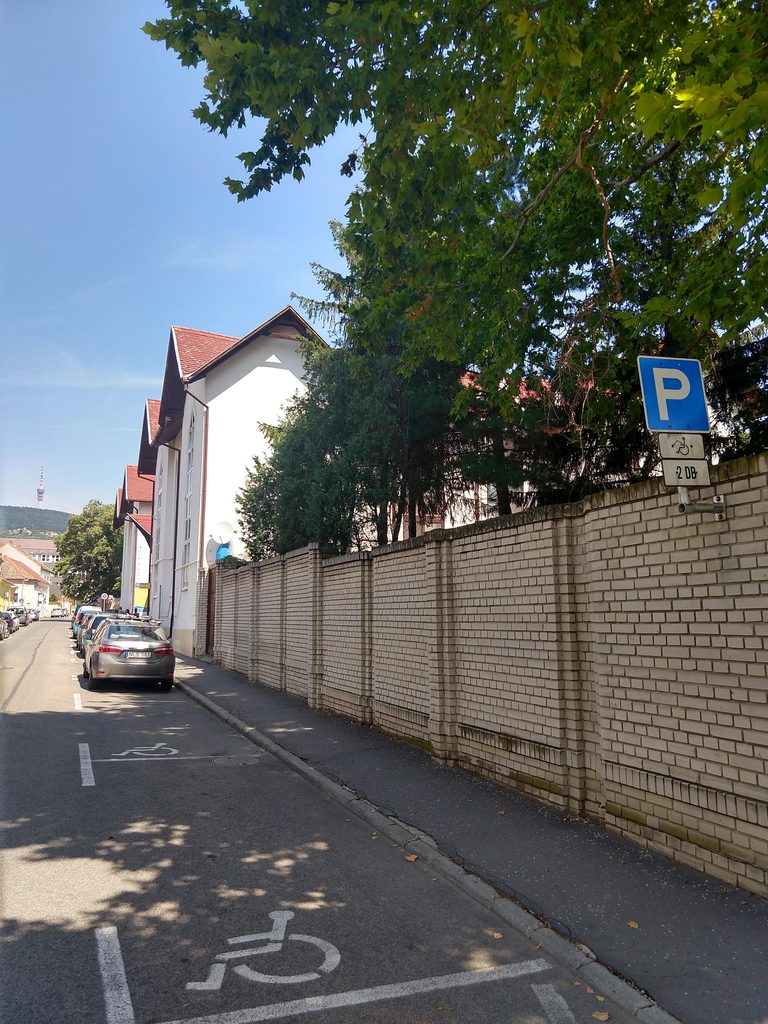 A hullámfürdőhöz vezető út. Jobb oldalon járda, melynek szélén egy nagy téglafal fut végig. A járda másik oldalán mozgássérült parkoló található.