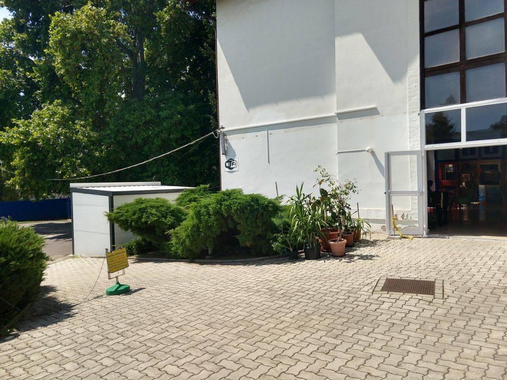 A hullámfürdő bejárata, az épülettel szemben. Nagy térkövezett út, zöld sövény, valamint cserepes növények találhatóak az épület mellett.