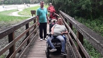Négy ember található a képen. Egy fa hídon mennek át. Közülük ketten kerekesszékesek. Tőlük jobbra van egy erdő, balra pedig egy parkoló.