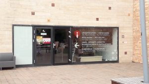 Zsolnay Kultúrális Központ információs pultjának bejárati üvegajtaja. A nagy üvegablakra az ajtó mellett a következők vannak felírva magyarul és angolul: Látogatóközpont, Pécsi galéria, Labor - interaktív varázstér, Planetárium.