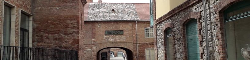 Épületek a Zsolnay negyedben. Bal oldalt tégla épület ablakokkal, előtte ablakok és kis terasz sötét korláttal szegélyezve. Középen egy köves út, távolabb egy boltíven megy át az épület alatt. Jobb oldalán a képen egy másik épület, ablakokkal és egy zöld faajtóval. A távolban két kémény látható.