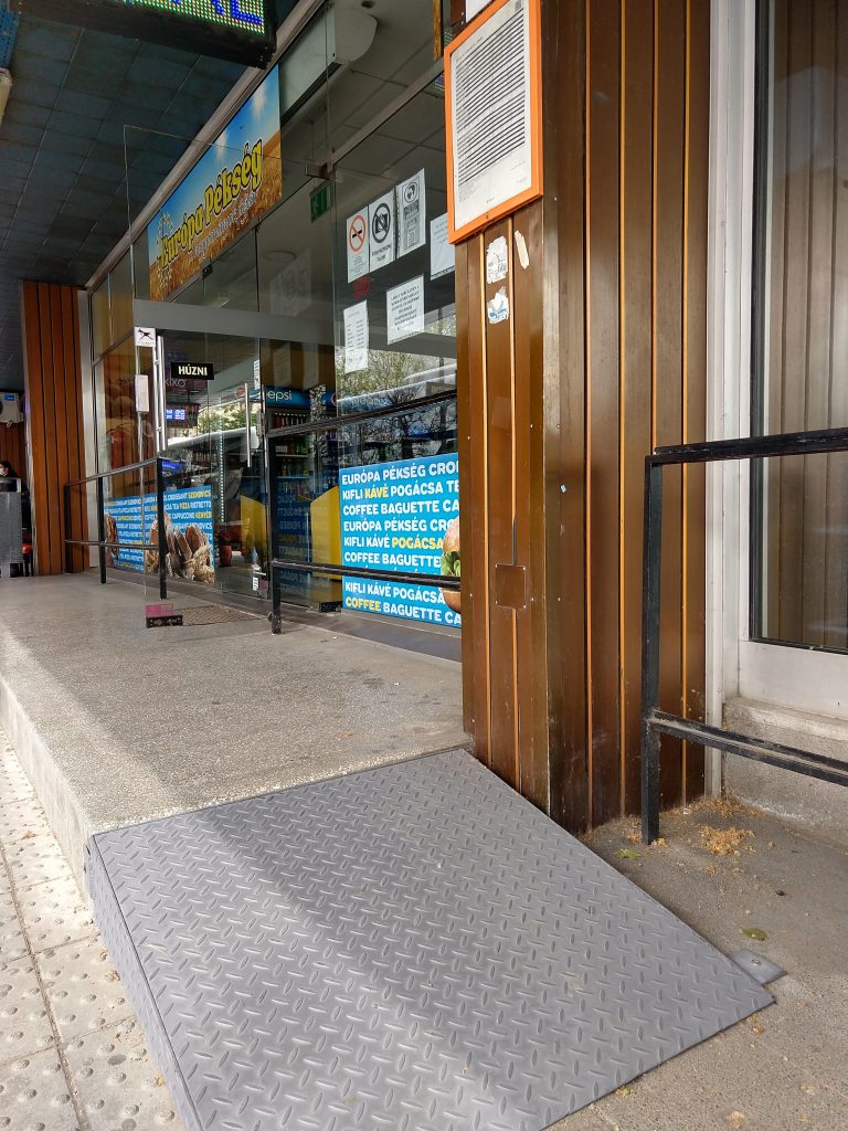 A pékség bejárata egy szinttel magasabban, egy lépcsőfokot egy hosszabb előtérre lépve érhető el. Oldalában rövid, alacsony dőlésszögű, állandó fém rámpa található.