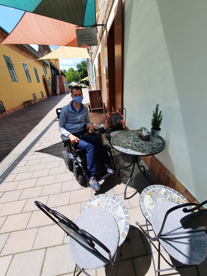 A boltok utcácskájában az árnyékban kisasztal és székek mellett pihen kerekesszékes tagunk, mögötte az utca folytatása látható.
