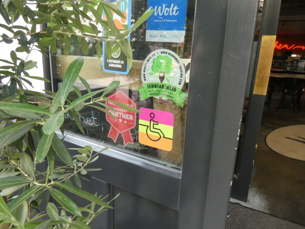 A szürke, üvegezett bejárati ajtón megtalálható a People First matricája, amely háttere három vízszintes sávban pink, zöld és narancssárga színű, és egy kerekesszékes személyt ábrázol.