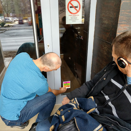 A Paulus étterem megkapja a People First akadálymentes matricáját az üvegajtójára. Egy kék pólós úriember helyezi fel az ajtóra, mellette egy kerekesszékes férfi figyeli őt.