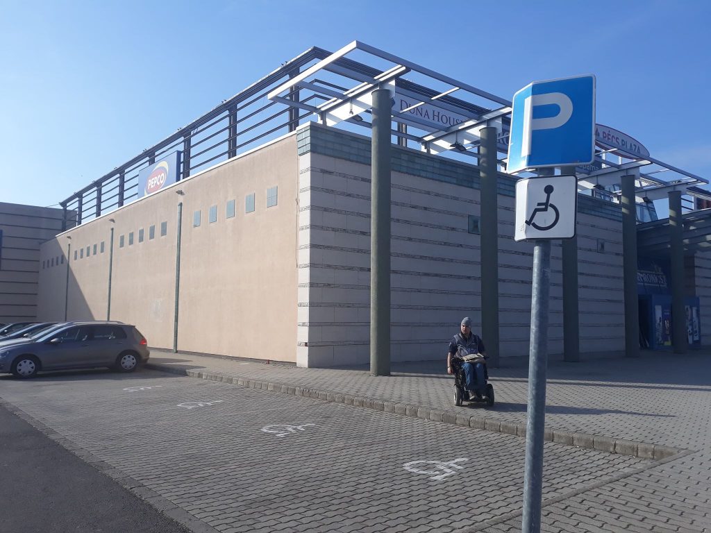 A képen a pláza látszik arról az oldalról, ahol a Pepco üzlethelység található. Van az épület mellett egy akadálymentes parkoló, ahol négy hely üresen van hagyva, amellett kocsik állnak. A parkoló fölött egy férfi van kerekesszékben.