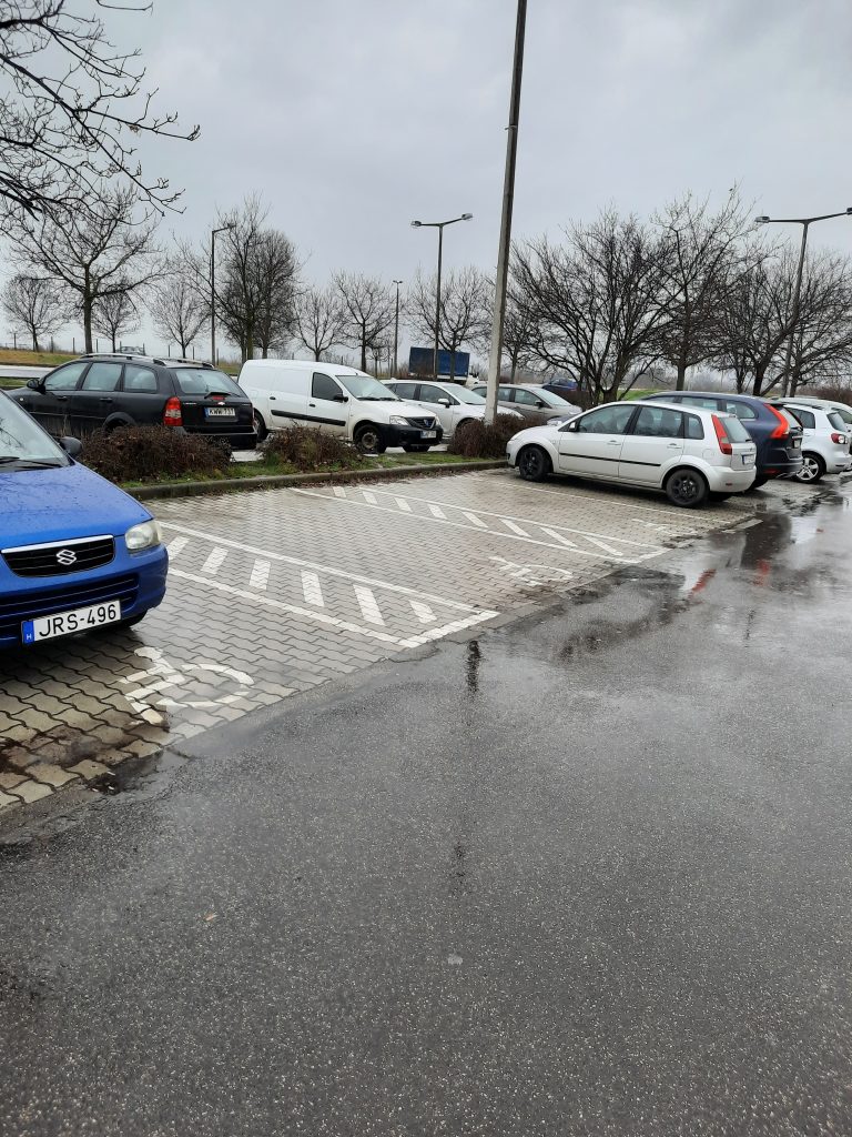 A képen a pláza parkolója van, ahol kocsik sorakoznak egymás mellett. Látható három akadálymentes parkoló, amik el vannak csíkos sávokkal választva egymástól. A bal szélső akadálymentes parkolóhelyen egy kék színű kocsi áll.