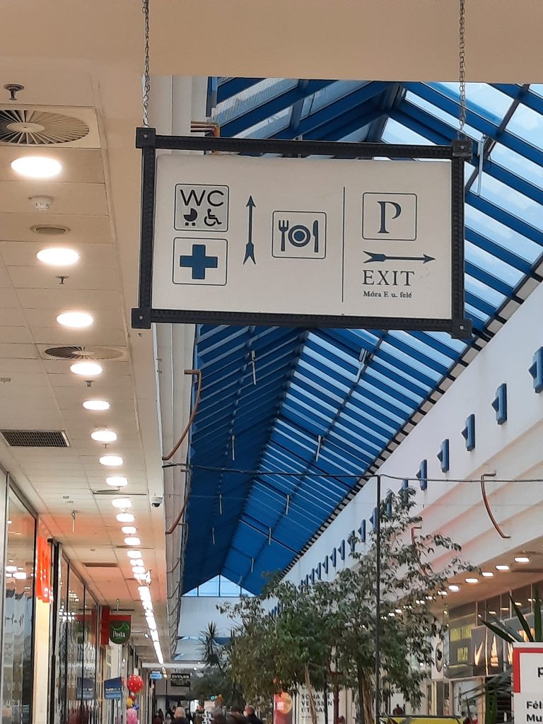 A Plázában a plafonról lóg le egy iránymutató tábla, kék színű szimbólumokkal és feliratokkal. A jobb oldalra mutató nyíl alatt EXIT felirat, fölötte parkolás jelző. Az előre mutató nyíl az éttermekhez, elsősegély szobához, illetve akadálymentes és babakocsis mosdókhoz irányít. A képen van még a pláza plafonja, illetve a plázában lévő növények.