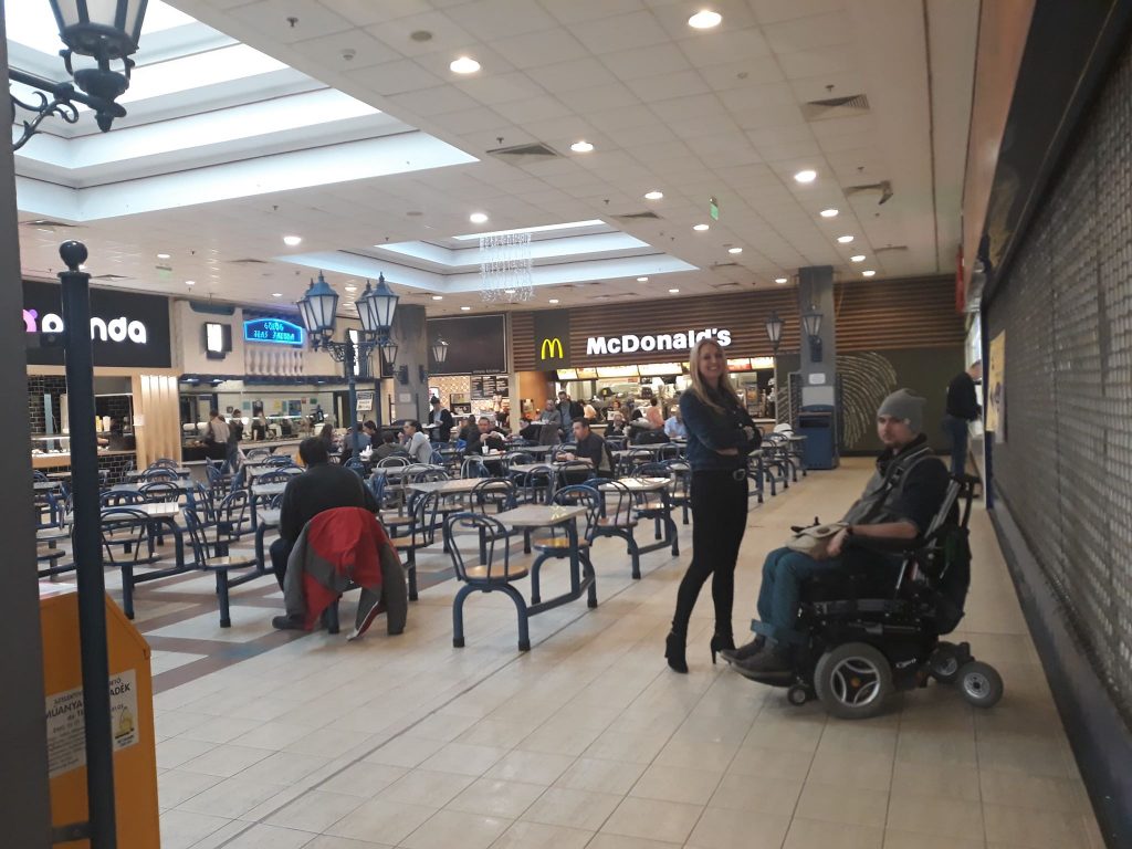 A képen a pláza ételudvara van, olyan asztalokkal, melyek a székekkel egybe vannak forrasztva, nem mozdítható. Vannak kék színű lámpaoszlopok a képen, illetve látszódik a McDonalds, a Purple Panda, emberek, ahogy esznek az asztaloknál, illetve a kép jobb oldalán a folyosón egy kerekesszékes férfi, mellette egy szőke hölgy áll.