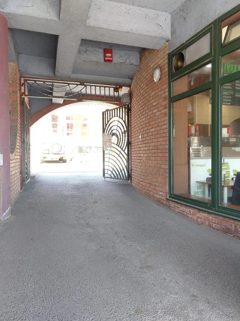 A Sopianae hotel autóbejárója egy enyhe emelkedő, a bejárata egy zöld színű nagykapu, amely a képen nyitva van. A bejárót téglafal és egy üveg kirakat öleli körbe.