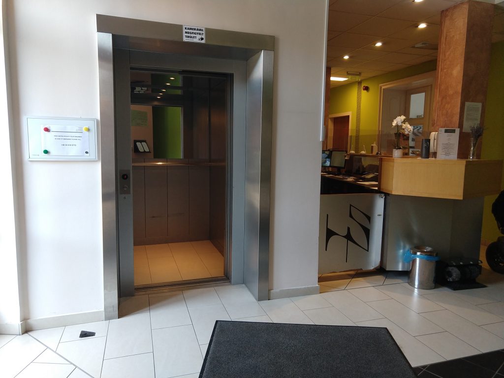Itt a Sopianae hotel liftje látható, tőle jobbra a hotel pultja található. A lift előtt sötétszürke szőnyeg pihen. A falak a liftnél fehérek, a pult előtt zöld színűek. A járólap fehér.