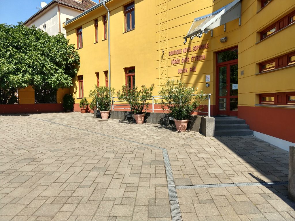 A Sopianae hotel udvara. Térkövezett. Elől lépcsőt látunk, akadálymentesítést, mozgássérült feljárót hátrébb találunk. Az épület maga sárga színt visel, az udvart virágzó leanderek ékesítik.