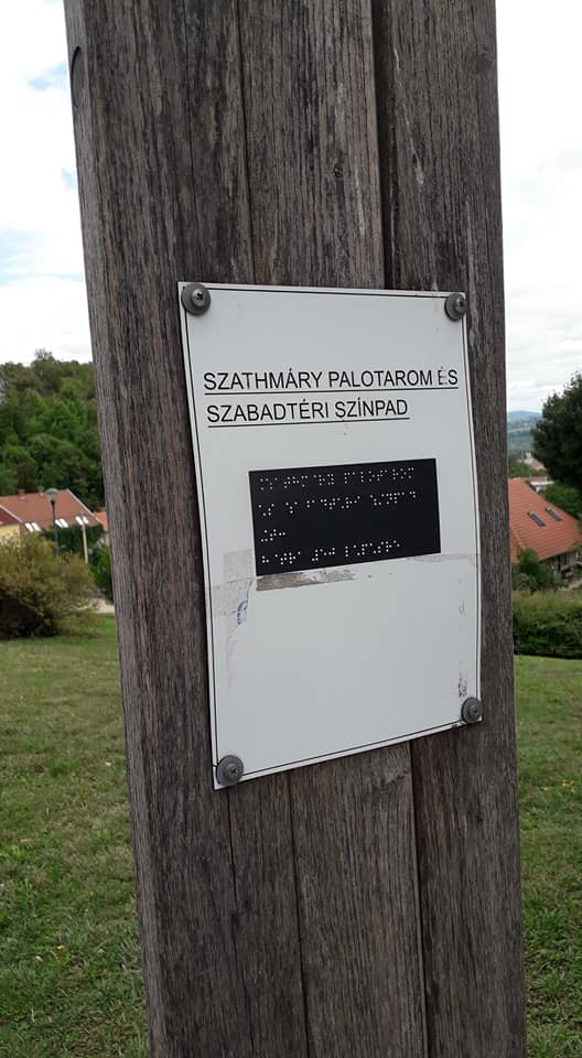 A Tettye túrán készült képen egy állított faléc látható, rajta egy négy sarkán felszegecselt műanyag fehér lap, melyre rá van írva fekete latin betűkkel és Braille írással ez a felirat: "Szathmáry palotarom és szabadtéri színpad".