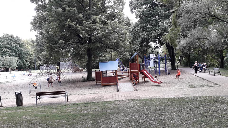 A Tettye téri játszótér kavicsos, van rajta többféle mászóka, az egyiken két gyerek mászik, mellettük egy felnőtt, egy piros csúszda, amin épp egy kislány csúszik, mérleghinta, forgó, amibe egy kisfiú kapaszkodik. A játszótér mentén ki van kövezve egy út, a szélén padokkal és kukákkal. Az egyik padon felnőttek ülnek a játszótérre nézve.