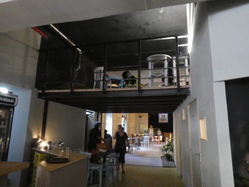 Két belső tér közötti folyosó. A folyosó padlója sárga, falai fehérek. A folyosón jobbra pult található, rajta poharakkal, mögötte pedig hűtővel. A pult után műanyag székek és egy asztal található. A folyosón jobbra 3 ajtó látható. A folyosó fölött egy másik terem részletét látjuk, itt is székek és asztalok vannak. A terem falai fekete színűek. A folyosó után szürke rámpa vezet egy nagyobb terembe, a rámpa mellett kövek vannak, rajtuk egy növény. A rámpa utáni teremben jobbra műanyag székek és egy faasztal található. Utána egy magas lámpa, velünk ellenkező irányba néz. Mellette egy üvegajtó látható. Utána még több asztal és szék, majd szemben egy nagy, fekete fal, előtte egy pulttal és rajta egy mikrofonnal. A fekete falon egy tábla áll, rajta a következő felirattal: SZABAD KIKÖTŐ. A felirat fölött egy logó látható: egy egyenes vonal, fölötte pedig egy hullámos vonal. A pult mellett, mindkét oldalt növények találhatók. Jobbra egy faasztal látható, közvetlenül a fal mellett. Mellette egy fehér kisasztal, rajta egy növénnyel és egy tükörrel.