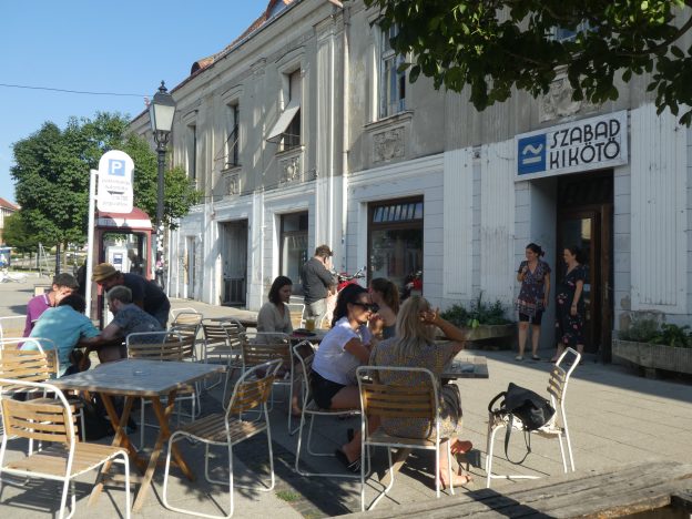 Előttünk faasztalok és székek láthatók, minden négyzet asztal körül négy szék. Összesen 6 ilyen szett van elhelyezve, háromnál ülnek is mások. Jobb oldalon egy alul fehérre festett, fölül szürkére festett épület, magas ablakokkal. Az ablakok között viszonylag nagy hely van kihagyva. Az épület jobb oldali szélén bejárati ajtó látható, fölötte táblával. A táblán a következő felirat áll: SZABAD KIKÖTŐ. A felirattól balra egy logó is látható: alul egy egyenes vonal, fölötte egy hullámos vonal.