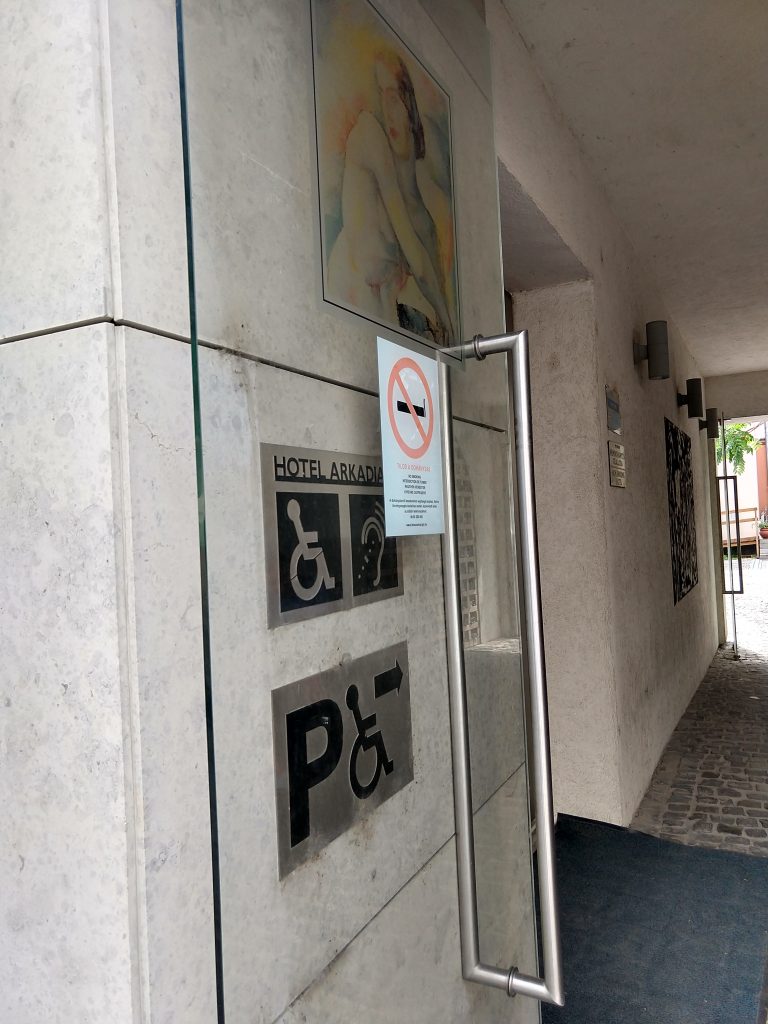 Bejárat, a falon két tábla. A fölsőn egy mozgáskorlátozott és egy hallássérült jele, azt jelzi, hogy ezek a személyek számára is akadálymentesített. Az alsó tábla a mozgáskorlátozott parkoló irányát mutatja. A táblák mellett egy dohányzást tiltó tábla is van, fölötte egy festménnyel.