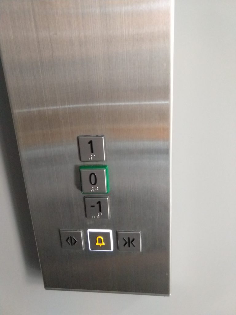 Lift kezelőpanelje. Fémes panelen találhatók nyomógombok. Egymás alatt, "1", "0" és "-1" feliratú gombok, mindegyiken Braille írás is látható. A "0" nyomógomb zöld kerettel is rendelkezik. Ezek a gombok alatt, egy sorban még három gomb. Az elsőn egy csíktól vezet kifele két nyíl (ez a liftajtó nyitására alkalmas), mellette egy csengő, fehér kerettel (ez a vészjelző), mellette egy csík felé vezető két nyíl (ez a liftajtó csukására alkalmas).
