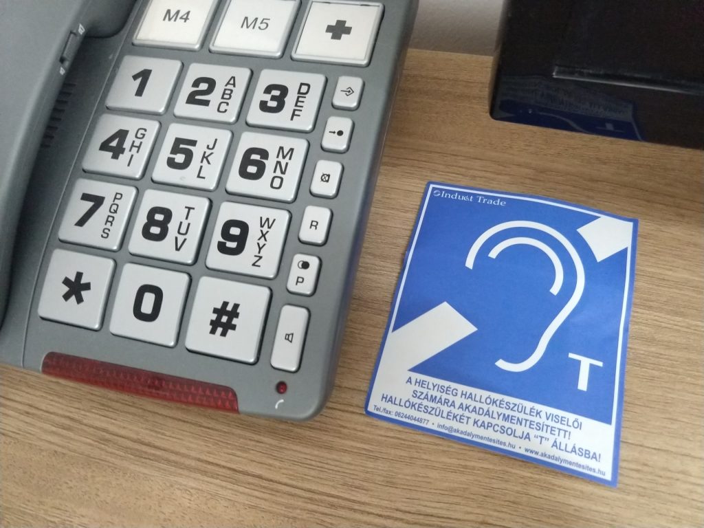 Asztal, melyen kagylós telefon és egy papír található. A telefon szürke színű, rajta a gombok fehérek. Az alap gombokon kívül látható még rajta egy M4 és egy M5 feliratú, és egy "+" jelzésű gomb is. A telefon mellett kék papír, melyen a következő felirat áll: A HELYISÉG HALLÓKÉSZÜLÉK VISELŐI SZÁMÁRA AKADÁLYMENTESÍTETT! HALLÓKÉSZÜLÉKÉT KAPCSOLJA "T" ÁLLÁSBA!